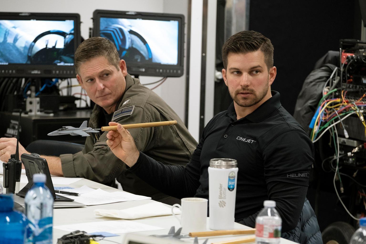 Kevin in a black CINEJET polo holding a jet model on a stick, seated beside Tom Cruise in a flight suit